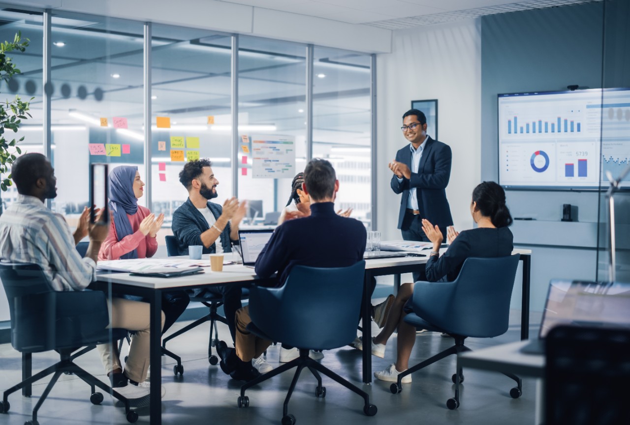 Multi-Ethnic Office Conference Room. Indian CEO does Presentation for Diverse Young Entrepreneurs, Talking, Using TV with Infographics, Statistics, Graphs. Businesspeople Invest in e-Commerce Startup