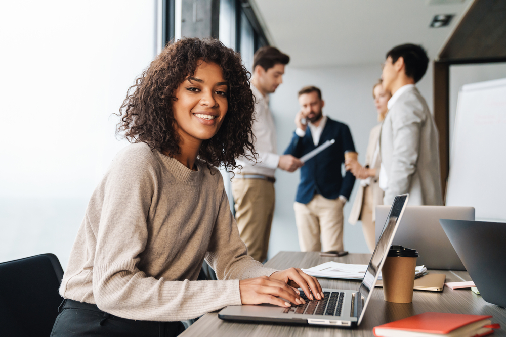 Attractive african young confident businesswoman sitting at the office table with group of colleagues in the background, working on laptop computer